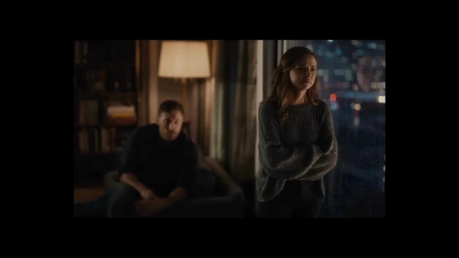 Sora 2 generated night scene of a woman standing with arms crossed beside a seated man in a lamp-lit apartment with city lights outside